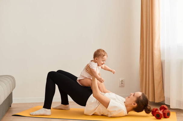 Side view portrait of happy woman wearing white t shirt and black leggins doing sport exercises with baby in hands, lying on yoga mat and doing workout for butt. - Foto, immagini