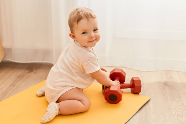 Horizontal shot of charming cute tiny infant kid sitting on yoga mat with dumbbells looking at camera with smile, exploring sport equipment. - Фото, изображение