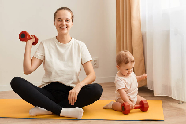 Portrait of adorable athletic woman wearing white t shirt and black leggins doing sport exercises with baby nearby, raising her arm with dumbbell, having workout for biceps. - Foto, Imagem