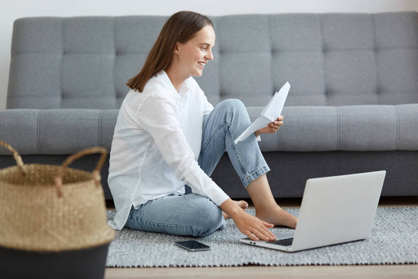 Εσωτερική λήψη χαμογελαστή μελαχρινή γυναίκα φορώντας λευκό πουκάμισο και τζιν κάθεται στο πάτωμα κοντά στον καναπέ, χρησιμοποιώντας φορητό υπολογιστή και κρατώντας έγγραφο χαρτί, πληρώνοντας κοινωφελείς υπηρεσίες. - Φωτογραφία, εικόνα