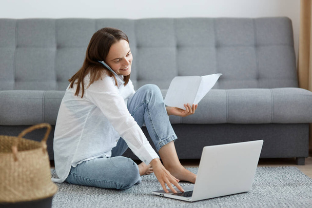 Indoor shot of joyful woman wearing white shirt and jeans sitting on floor near sofa, using laptop and talking phone, expressing happiness, working and having pleasant conversation. - Valokuva, kuva
