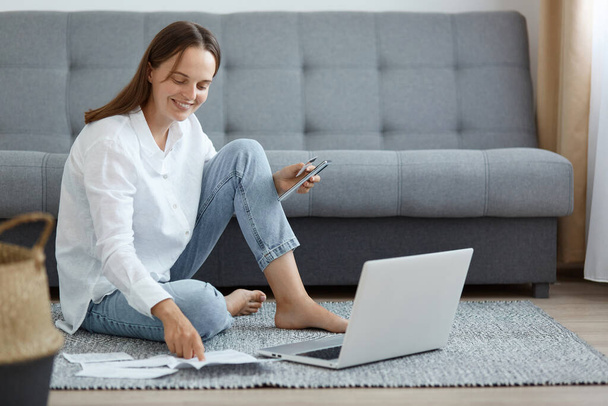 Indoor shot of smiling woman wearing white shirt and jeans sitting on floor near sofa, using laptop, holding credit card, checking information in utilities bills on paper. - Фото, изображение