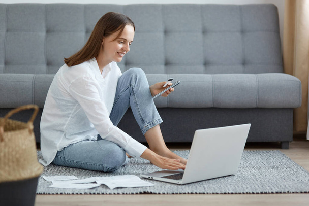Image of winsome dark haired woman wearing white shirt and jeans sitting on floor near sofa, using laptop, holding credit card, soving problems with bills, online payment. - Photo, Image