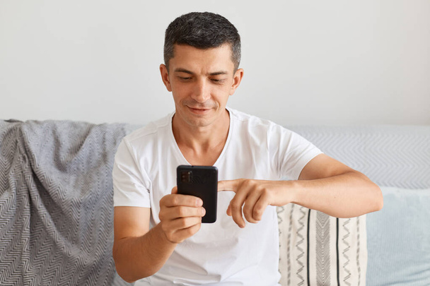 Smiling handsome young adult man wearing white t shirt sitting on sofa at home, using cell phone for checking social networks or email, looking at device screen. - Foto, imagen