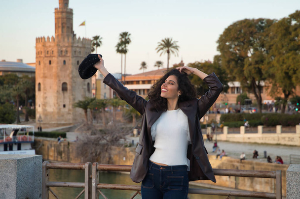 Young and beautiful woman, brunette, with curly hair, with leather jacket and white shirt with a beret in her hand, smiling happily, leaning on the railing of a bridge. Concept holidays, travel. - Photo, Image