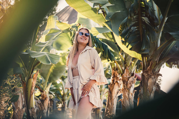 A beautiful young woman with long blond hair of European appearance stands near the banana trees. Girl in tropical forest on a sunny summer day - Foto, afbeelding