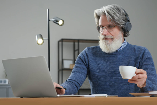 Middle aged man with laptop, headphones and cup of drink learning at table indoors - Photo, image