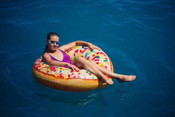 Een jong mooi meisje in een helder zwempak ligt op een grote opblaasbare ring en drijft op de blauwe zee op een zonnige zomerdag - Foto, afbeelding