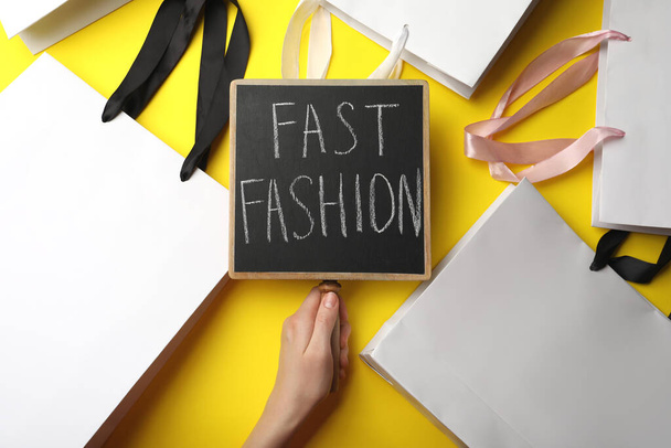 Woman holding small chalkboard with phrase Fast Fashion and white shopping bags on yellow background, top view - Photo, Image