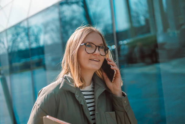 Retrato de sonrisa Mujer de negocios rubia europea de 30 años usando anteojos hablando con teléfono cerca del edificio de oficinas exteriores - Foto, imagen