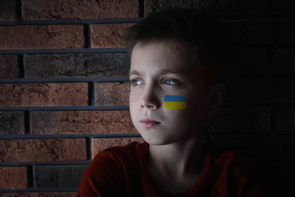 Sad little boy with picture of Ukrainian flag on cheek near brick wall. Stop war in Ukraine - 写真・画像