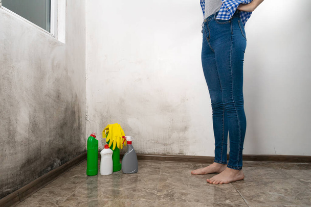 Nahaufnahme schlanker weiblicher Beine in Jeans, die in der Nähe einer Wand mit schwarzem Schimmel mit Reinigungsmitteln stehen. Konzept der Reinigung, Hausarbeit - Foto, Bild