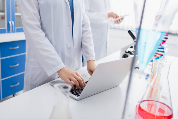 cropped view of scientist typing on laptop in laboratory near blurred colleague - Foto, afbeelding