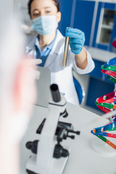 Tıbbi maskeli bulanık genetikçi mikroskop ve DNA modelinin yanında test tüpü tutuyor. - Fotoğraf, Görsel
