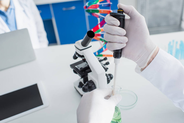 обрезанный вид биотехнолога в латексных перчатках с микропипеткой и фляжкой рядом с микроскопом - Фото, изображение