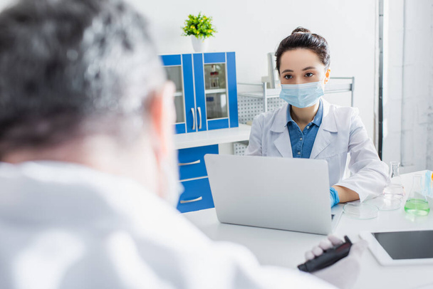 young scientist in medical mask working on laptop near blurred colleague - Photo, Image