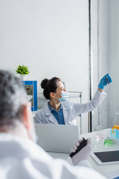Biotechnologe in medizinischer Maske hält Reagenzglas neben Laptop und verschwommenem Kollegen - Foto, Bild