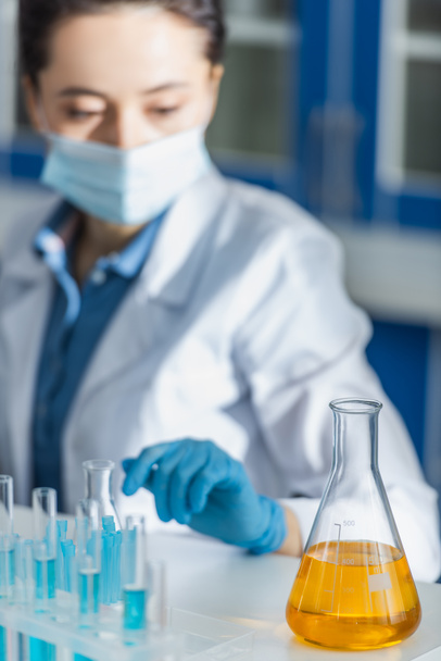 selective focus of flask with yellow liquid near blurred bioengineer in medical mask - Photo, image