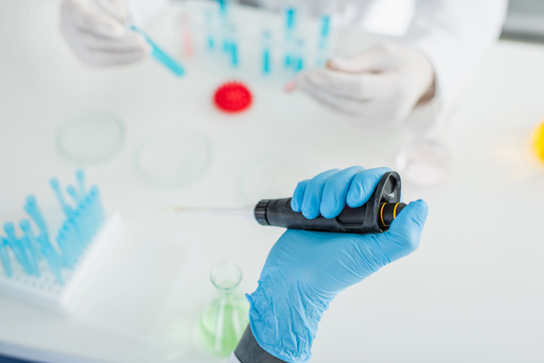 vue recadrée du scientifique dans un gant en latex tenant une micropipette près d'un collègue flou - Photo, image