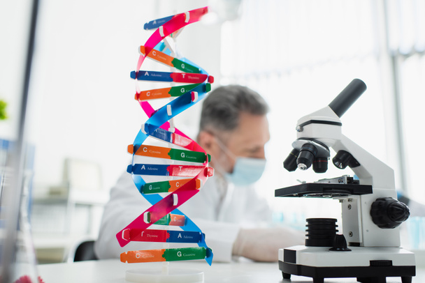 model DNA a mikroskop v blízkosti rozmazaného biotechnologa v lékařské masce na rozmazaném pozadí - Fotografie, Obrázek