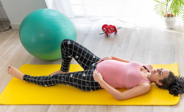 Genç, güzel, hamile bir kadın spor paspasına uzanıp bacak egzersizleri yapıyor ve sırtı geriniyor. Hamilelikte konsept sağlık ve spor - Fotoğraf, Görsel