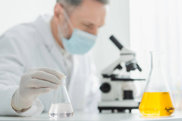 размытый биотехнолог в медицинской маске, смотрящий в микроскоп возле колб с жидкостью - Фото, изображение