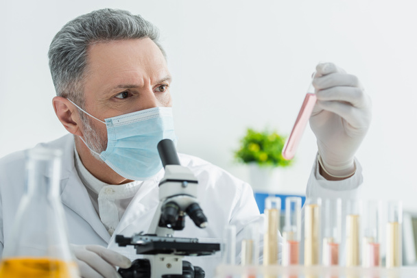 седовласый биоинженер в медицинской маске смотрит на пробирку возле микроскопа - Фото, изображение