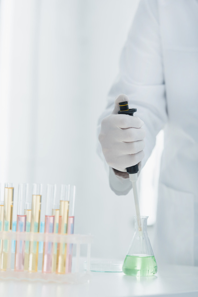 vista parcial del genetista en guante de látex sosteniendo micropipeta cerca del matraz y tubos de ensayo - Foto, Imagen