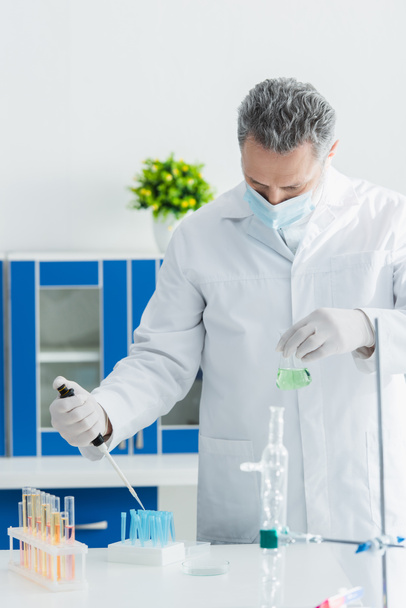 биоинженер в медицинской маске держит микропипетку и колбу во время работы с пробирками в лаборатории - Фото, изображение