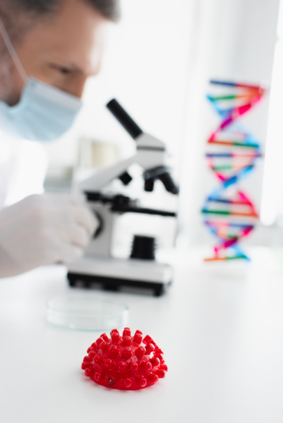 селективный фокус модели красных коронавирусов вблизи вирусолога в медицинской маске на размытом фоне - Фото, изображение