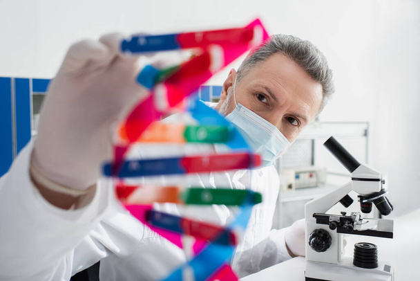 genetista en máscara médica cerca del modelo borroso de ADN y microscopio en laboratorio - Foto, Imagen