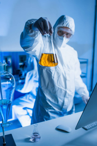 zamazany naukowiec w kolbie w kombinezonie ochronnym z pomarańczowym płynem w laboratorium - Zdjęcie, obraz