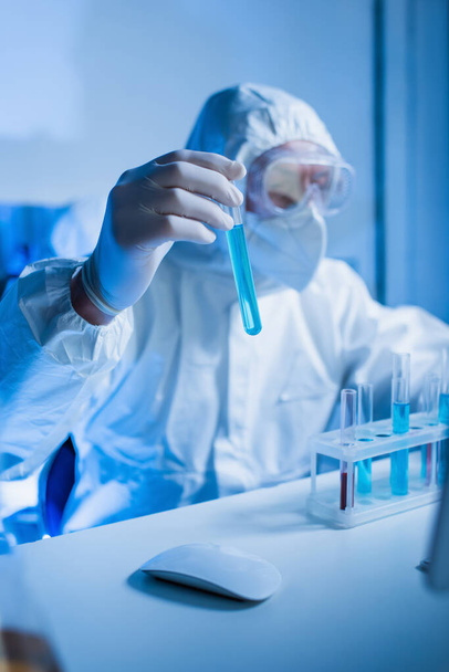blurred virologist in hazmat suit and goggles holding test tube with blue liquid - Valokuva, kuva