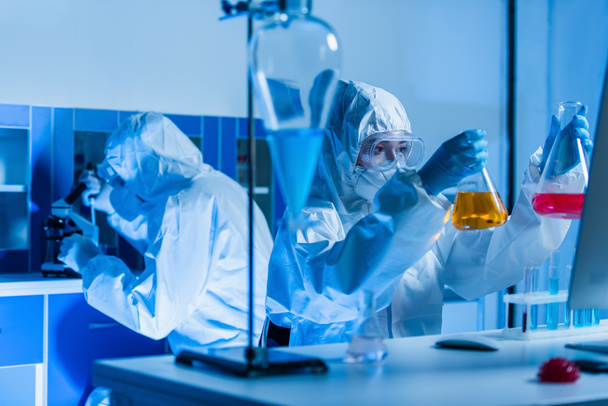 генетик в индивидуальном защитном оборудовании держит колбы с красочной жидкостью в лаборатории - Фото, изображение