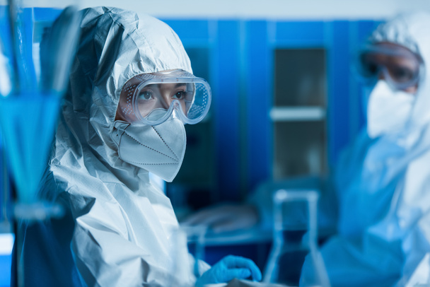 kobieta w goglach, maska medyczna i kombinezon ochronny w pobliżu naukowca na zamazanym tle - Zdjęcie, obraz