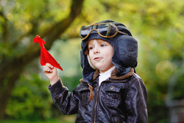 Niño feliz niño en casco piloto y uniforme jugando con el avión de juguete rojo. Sonriente niño preescolar soñando y divirtiéndose. Educación, profesión, concepto de sueño - Foto, imagen