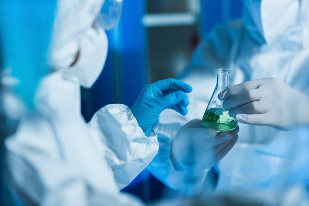 обрезанный вид биоинженера в латексных перчатках, держащего фляжку с жидкостью рядом с размытым ученым - Фото, изображение