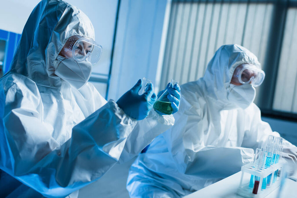 científico en traje de materiales peligrosos sosteniendo frascos médicos mientras trabaja con un colega en el laboratorio - Foto, imagen