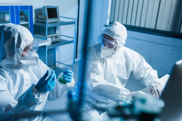 биоинженер с фляжками смотрит на коллегу в защитном костюме возле компьютера, размытый передний план - Фото, изображение