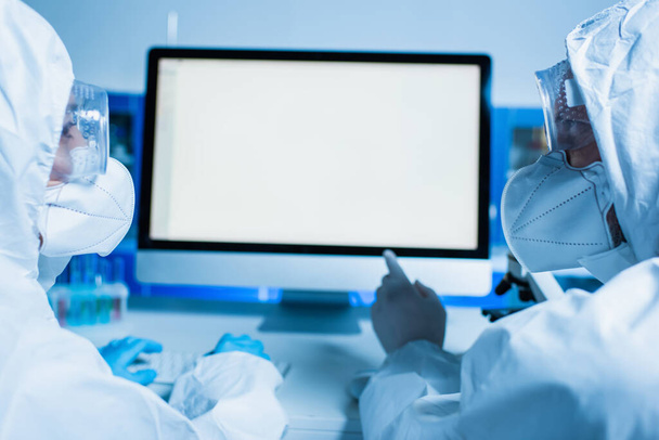 Tehlikeli madde giysisi ve tıbbi maskeli bilim adamı meslektaşıyla çalışırken monitöre beyaz ekranla bakıyor. - Fotoğraf, Görsel