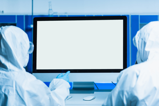 vista posteriore dello scienziato in tuta hazmat che punta al monitor con schermo bianco vicino al collega - Foto, immagini