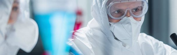 tudós vegyvédelmi ruha, szemüveg és orvosi maszk közelében elmosódott kolléga a laborban, banner - Fotó, kép