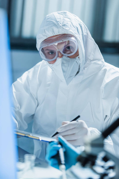 virólogo en traje de materiales peligrosos y gafas mirando a la cámara en primer plano borroso - Foto, Imagen