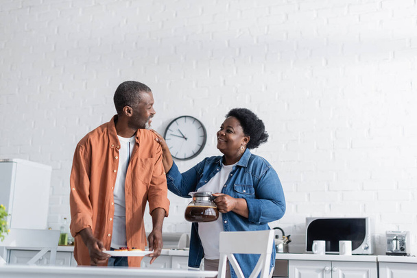 felice uomo africano americano che tiene frittelle sul piatto e guardando moglie con caffettiera - Foto, immagini