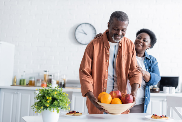 senior african american man putting bowl with fruits on breakfast table near smiling wife  - Foto, afbeelding