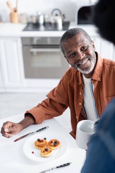 счастливый африканский американец смотрит на жену, держащую чашку во время завтрака  - Фото, изображение