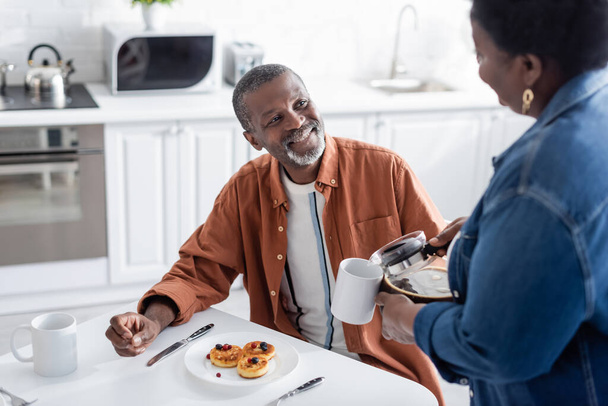 glücklich afrikanisch-amerikanischer Mann schaut Frau beim Frühstück beim Kaffeeservieren an  - Foto, Bild
