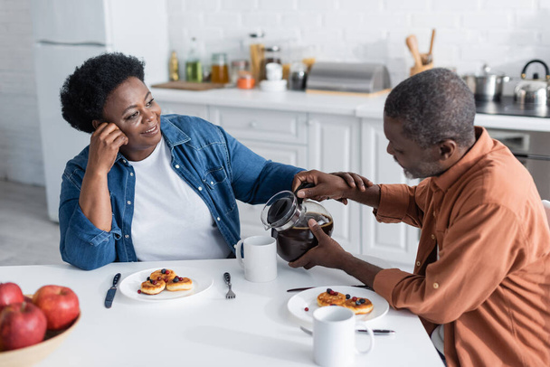 senior african american man pouring coffee to cheerful wife during breakfast  - Fotó, kép