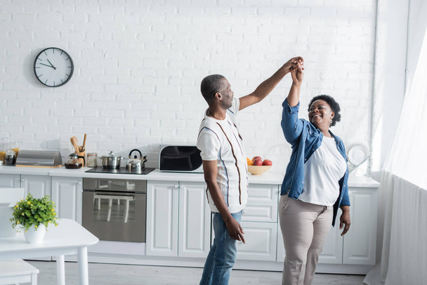retired african american couple smiling while dancing in kitchen  - Foto, Imagen