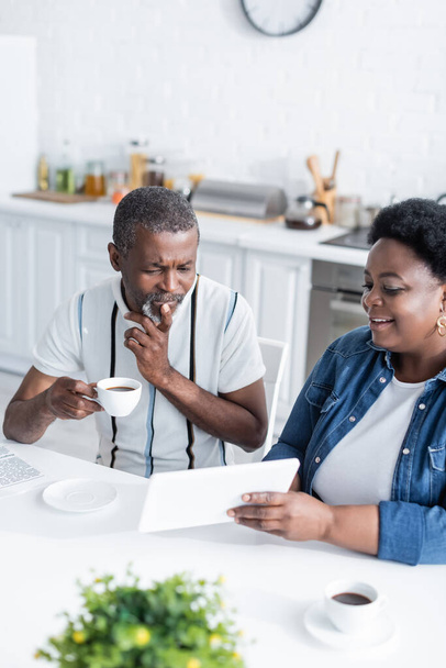 cheerful african american woman showing digital tablet to senior husband with cup of coffee  - Fotó, kép